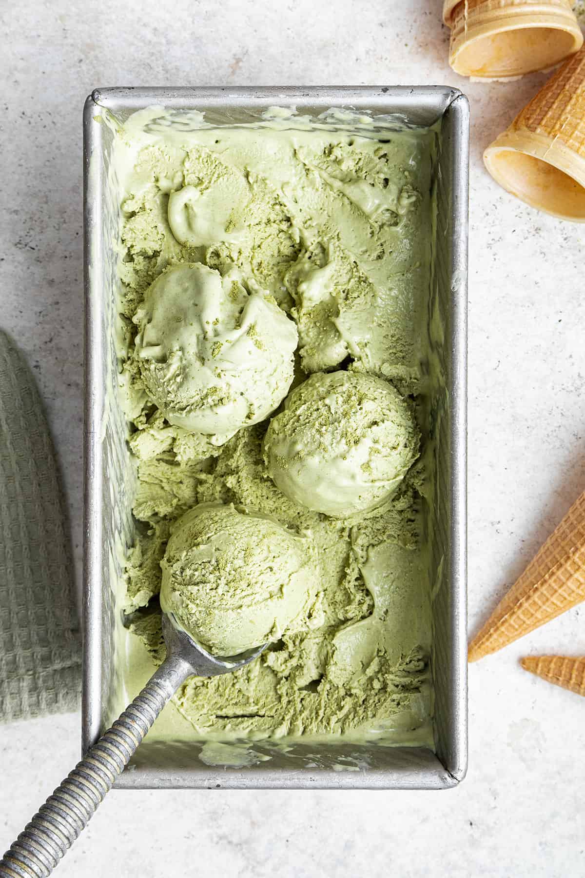 Overhead view of a loaf pan filled with matcha ice cream, with an ice cream scoop scooping a ball of ice cream
