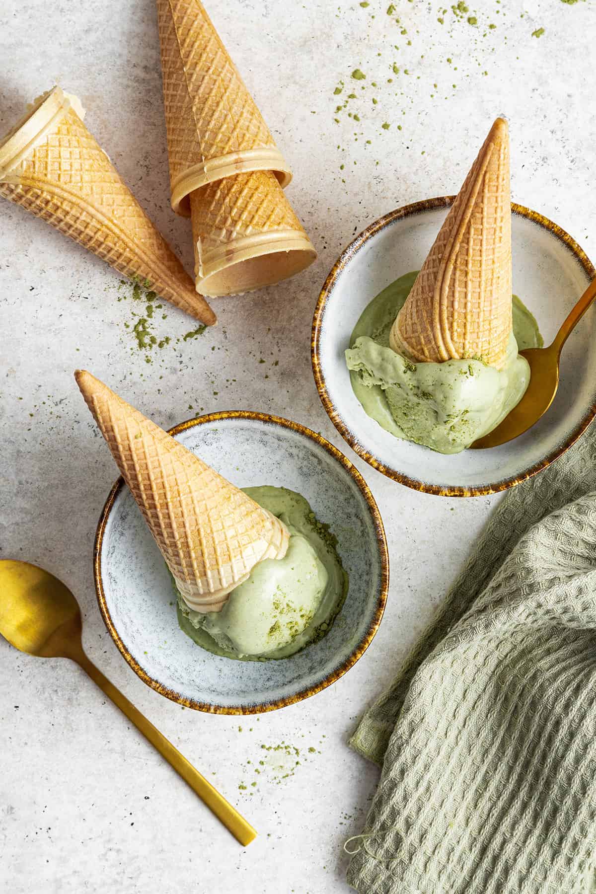 Two ice cream cones with matcha ice cream scoops, face down in bowls, next to a few cones, a spoon, and a kitchen towel