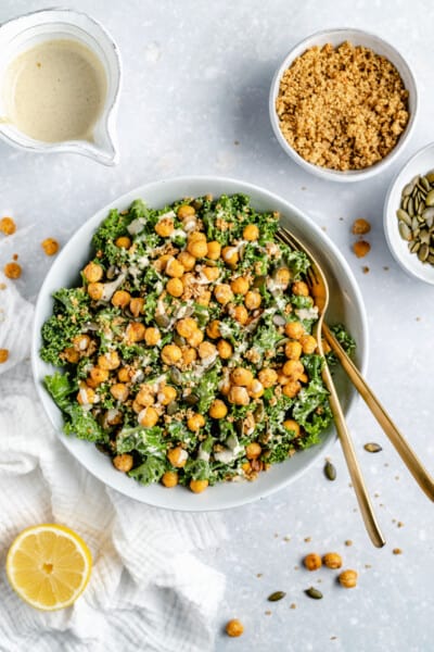 Overhead view of vegan kale Caesar salad in a white bowl topped with crispy chickpeas and tahini dressing, surrounded by salad ingredients.