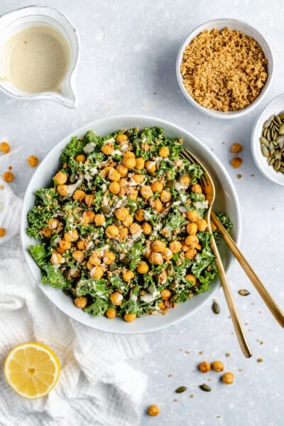 Overhead view of vegan kale Caesar salad in a white bowl topped with crispy chickpeas and tahini dressing, surrounded by salad ingredients.