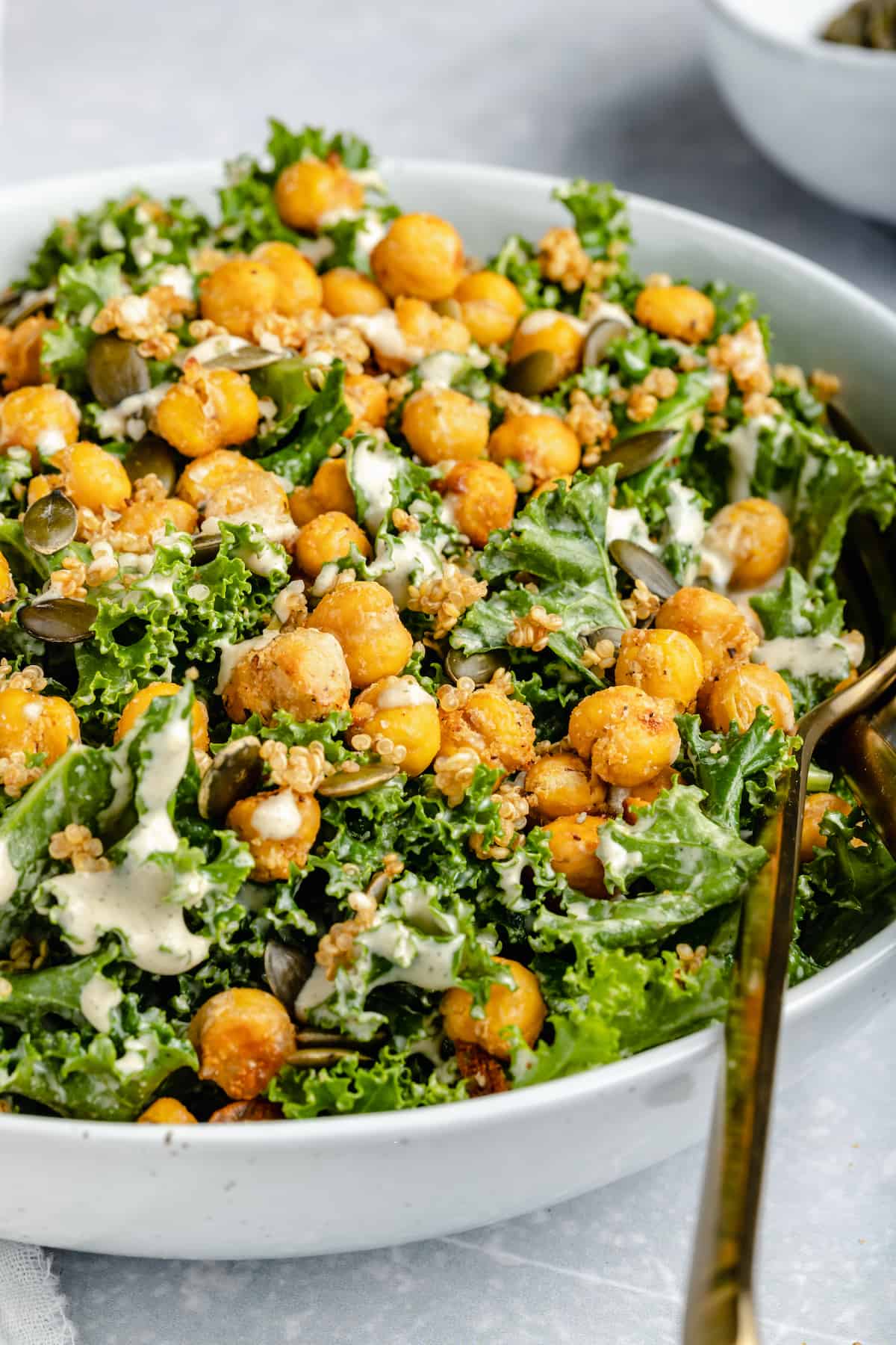 Vegan kale Caesar salad in a white bowl topped with crispy chickpeas and tahini dressing, with a spoon for serving.