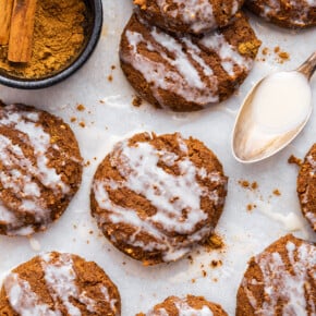 Overhead view of frosted pumpkin cookies on parchment paper with spoonful of coconut butter and bowl of cinnamon