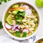 Overhead view of pozole verde in bowl, topped with sliced jalapeños, radishes, lime wedges, avocado, cilantro, and tortilla strips