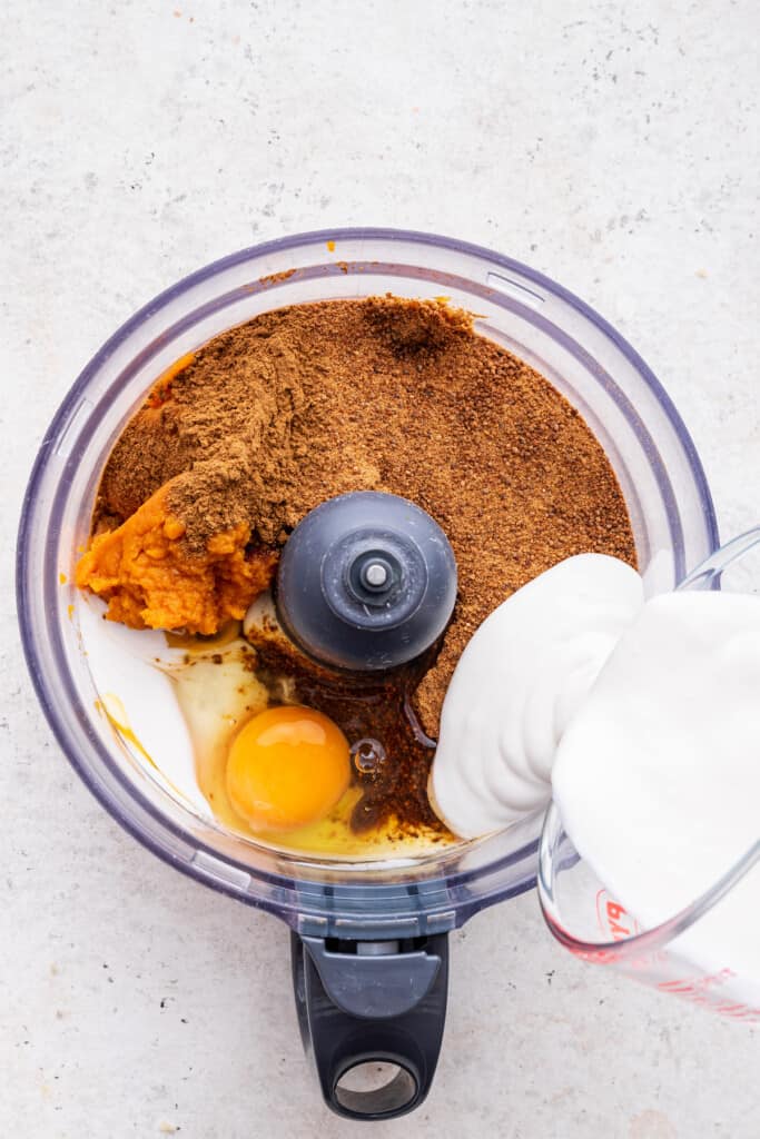 Overhead view of coconut milk being poured into food processor with other pumpkin pie ingredients