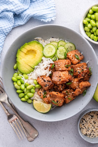 Overhead view of air fryer salmon bites in bowl with rice, cucumbers, edamame, avocado, and lime