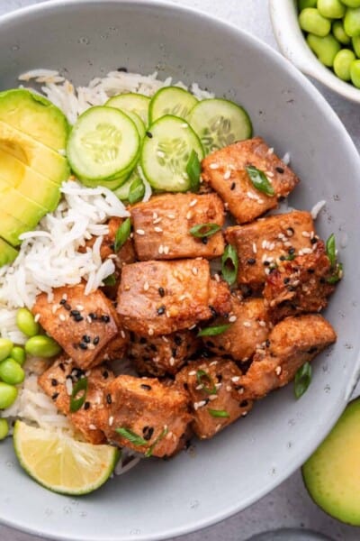Overhead view of air fryer salmon bites in bowl with rice, cucumbers, edamame, avocado, and lime