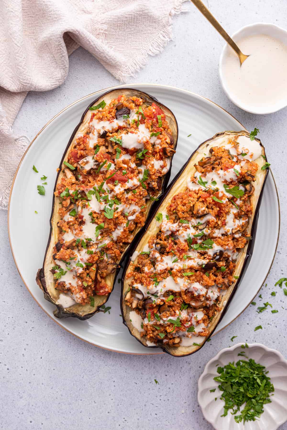 This Mediterranean-inspired stuffed eggplant recipe is full of flavor, packed with veggies and quinoa, and it takes just 30 minutes to make! Overhead view of stuffed eggplant on plate next to bowls of tahini and parsley