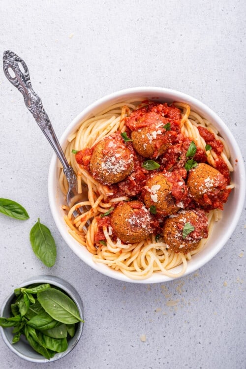 Overhead view of vegan meatballs in bowl of spaghetti set next to bowl of basil leaves