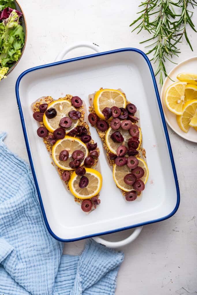 Overhead view of halibut topped with mustard, lemon slices, and olives