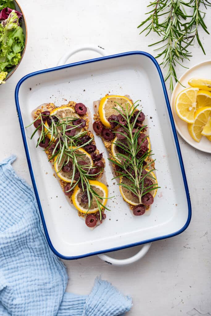 Overhead view of halibut topped with lemon slices, mustard, olives, and rosemary before baking