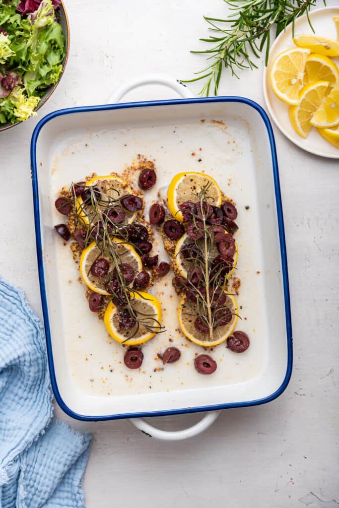 Overhead view of baked halibut in baking dish topped with lemon slices, olives, and rosemary sprigs