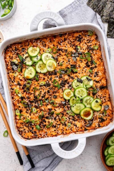 Overhead view of salmon sushi bake in baking dish with sliced cucumbers and green onions for garnish