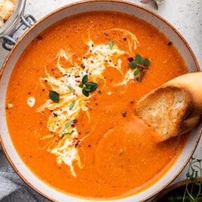 Bowl of roasted red pepper soup garnished with coconut milk, herbs, and bread