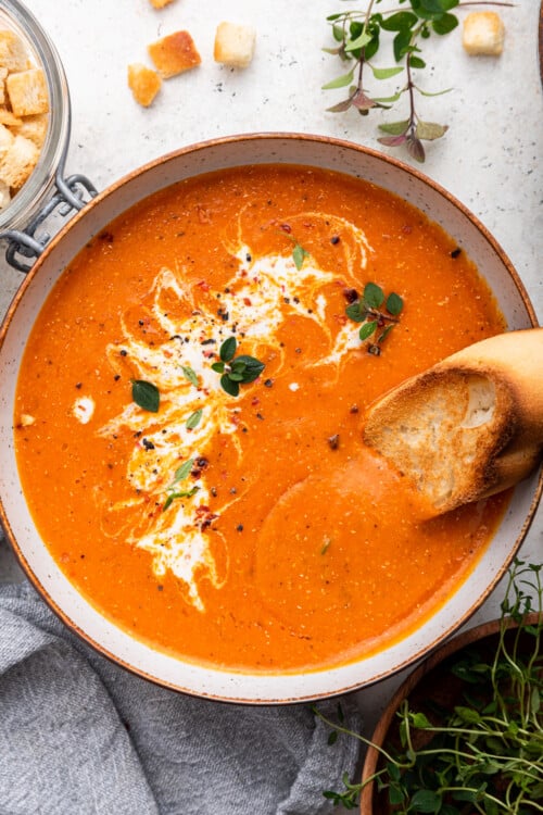 Bowl of roasted red pepper soup garnished with coconut milk, herbs, and bread