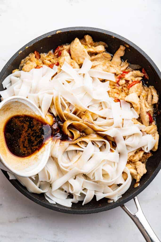 Pouring sauce into pan with rice noodles, chicken, and vegetables