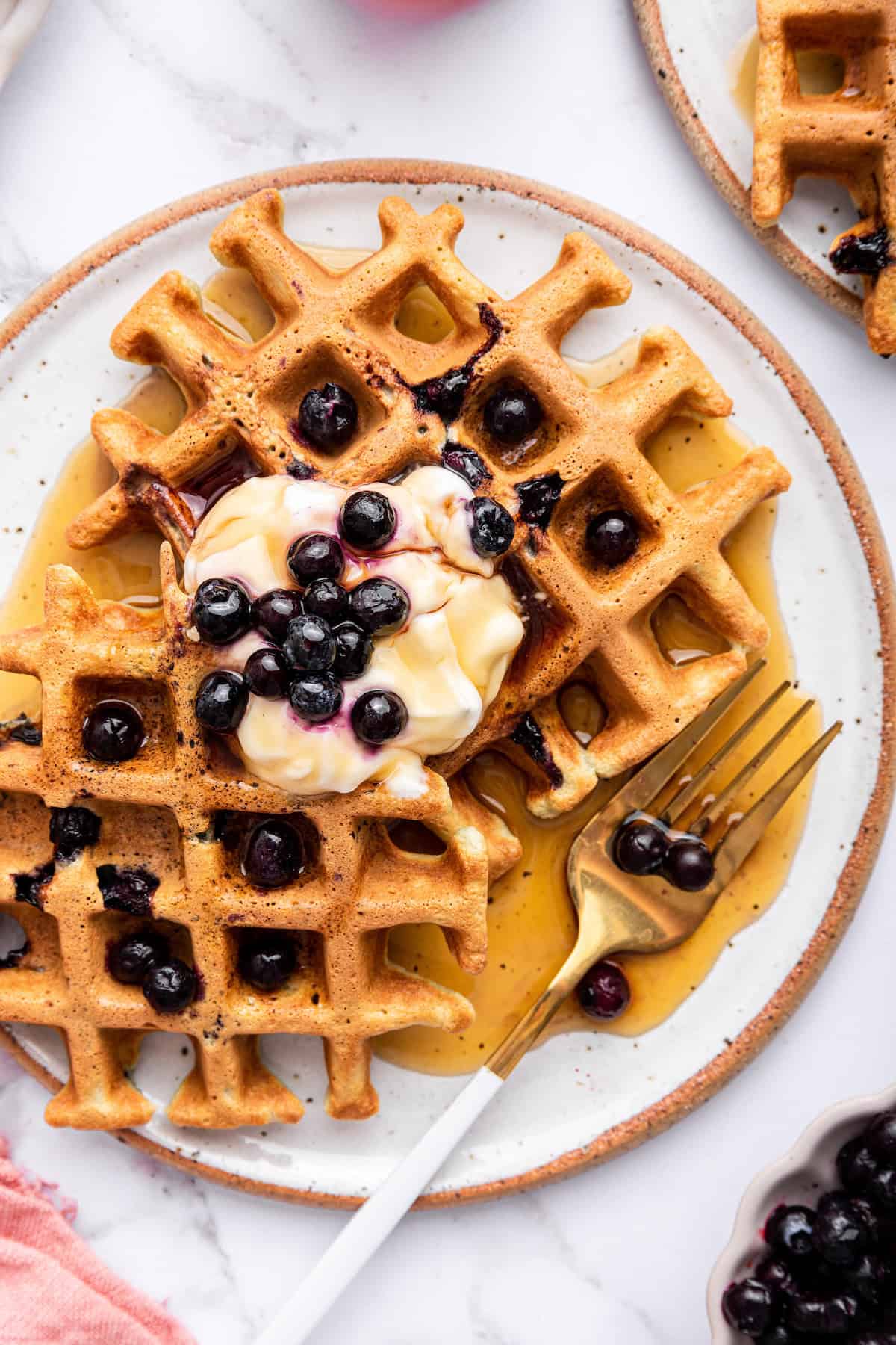 With their light, fluffy interior, crispy edges and loads of juicy berries, these easy homemade blueberry waffles are guaranteed to be a hit! Overhead view of 2 blueberry waffles on plate with maple syrup