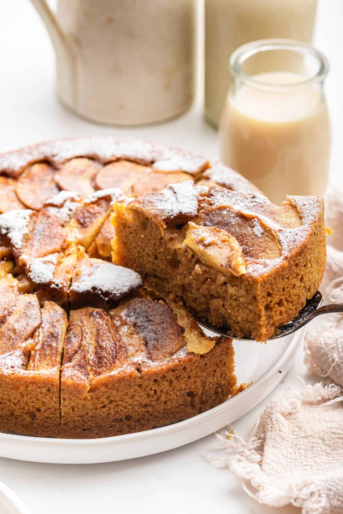 This German apple cake is super moist, topped with caramelized apples, and spiced with cozy cinnamon. The perfect cake to celebrate fall! Lifting slice of German apple cake from platter