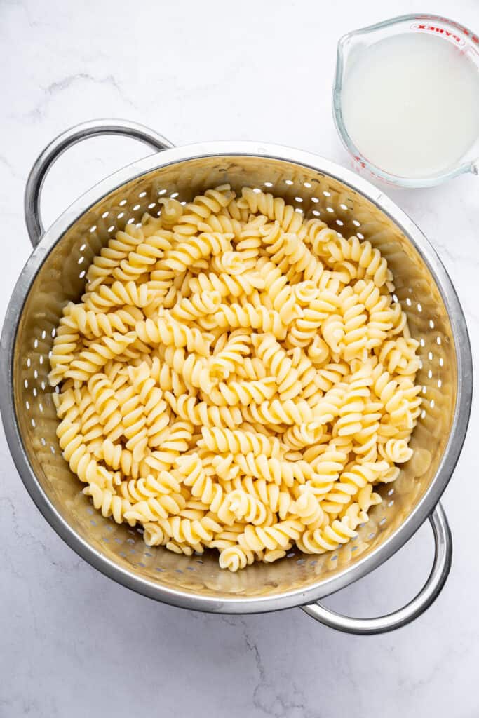 Drained rotini in colander with reserved cooking liquid in measuring cup