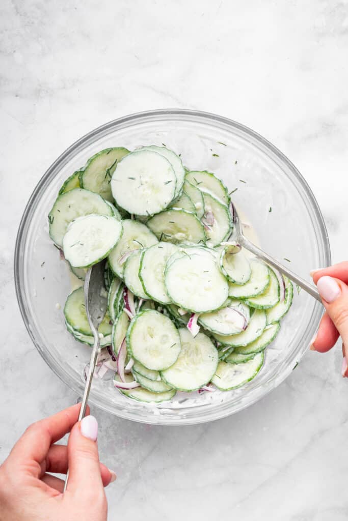 Tossing creamy cucumber salad in glass bowl with two spoons