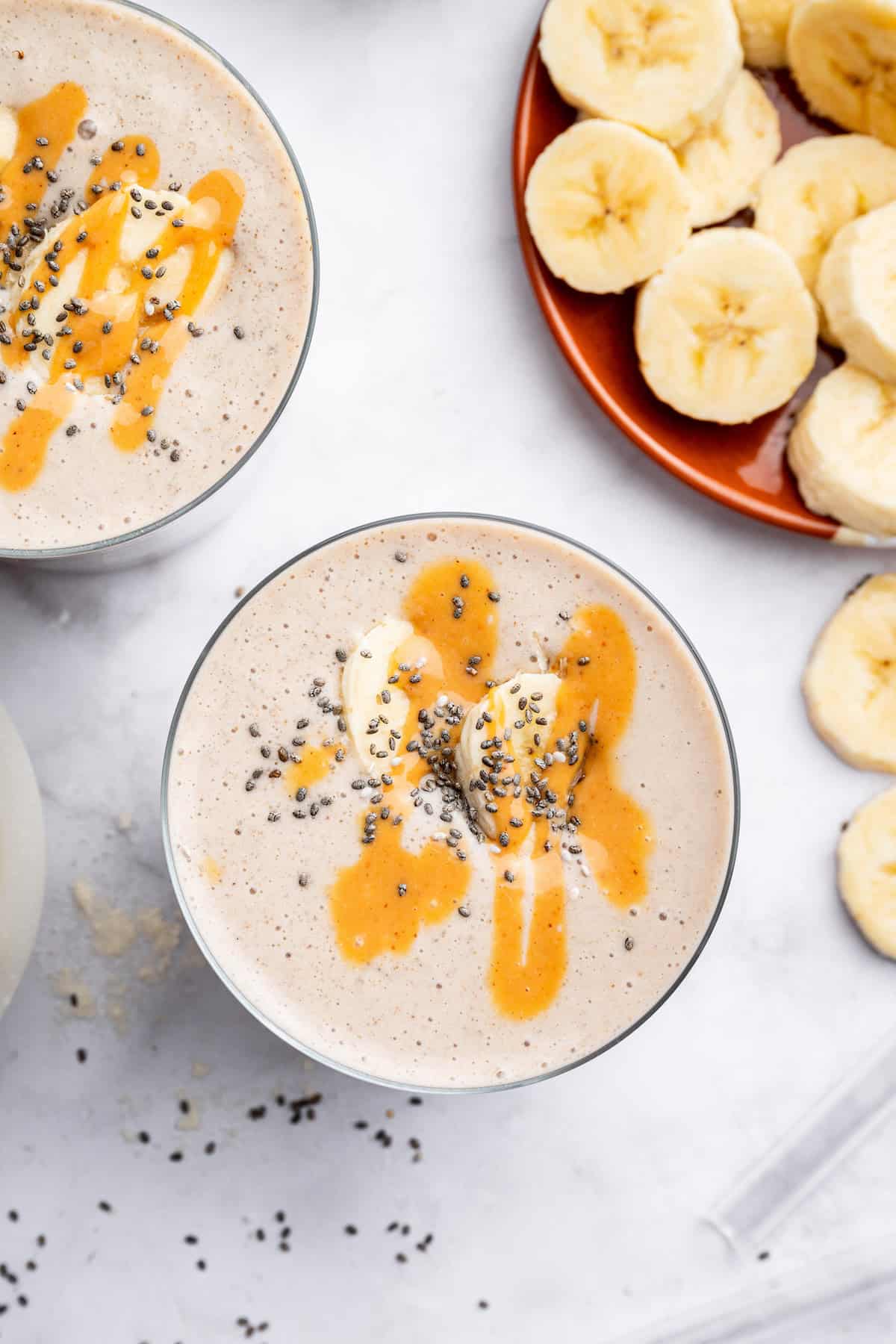 Overhead view of peanut butter banana smoothies in glasses with plate of sliced bananas