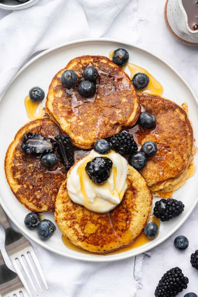 Cottage cheese pancakes are tender and fluffy, with more protein than a traditional pancake. Stash some in the freezer for easy breakfasts! Overhead view of cottage cheese pancakes on plate with berries, yogurt, and maple syrup