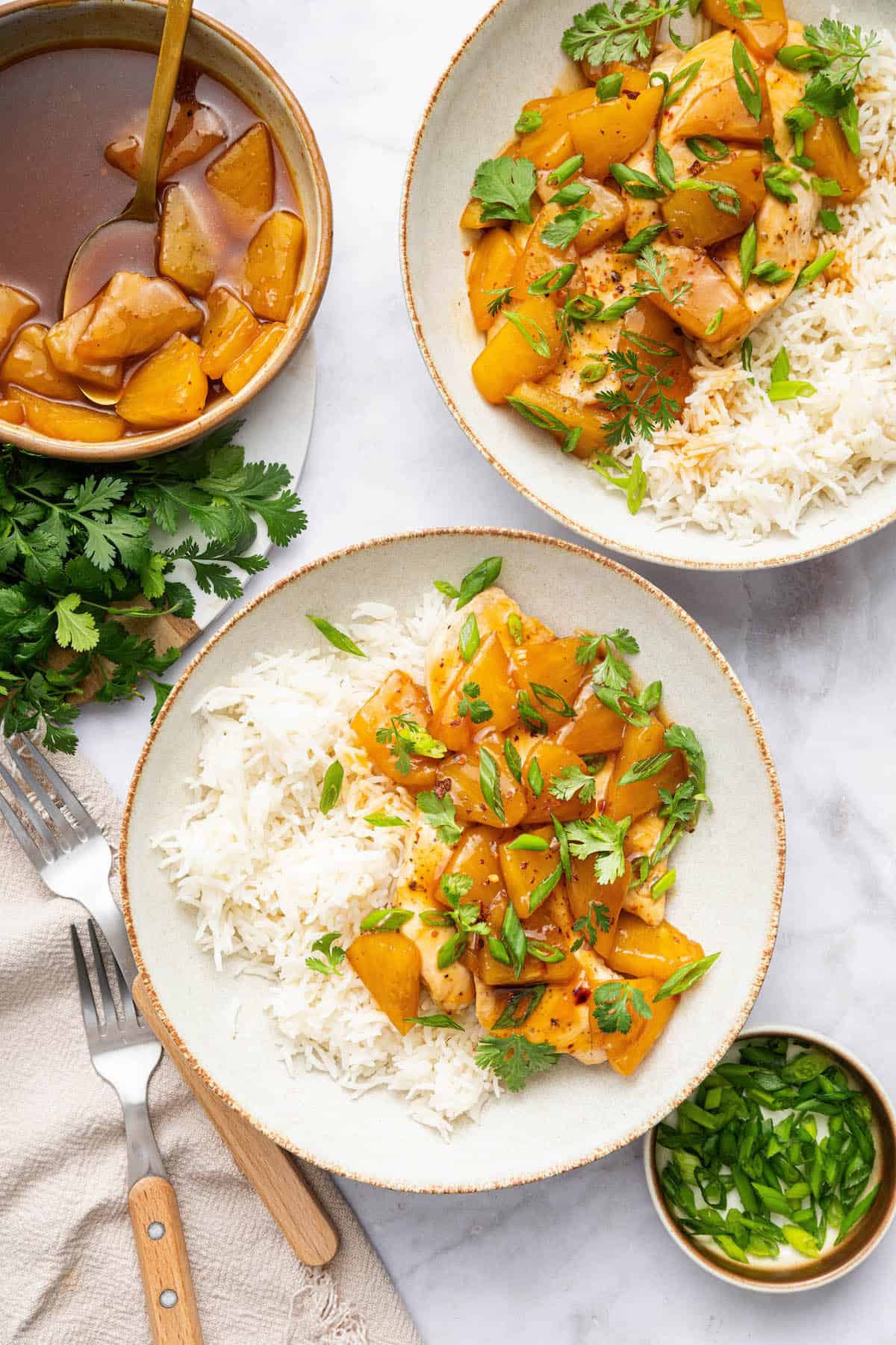 Two bowls of pineapple chicken with white rice with pan of sauce and pineapple