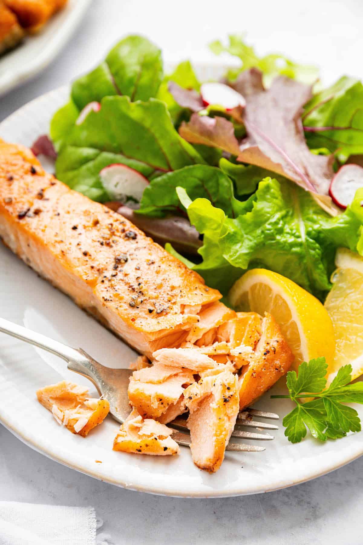 Yes, you can cook frozen salmon in the air fryer! It’s done in 25 minutes, ready for adding your favorite sauces, garnishes, and sides. Fork cutting into air fried frozen salmon on plate with salad