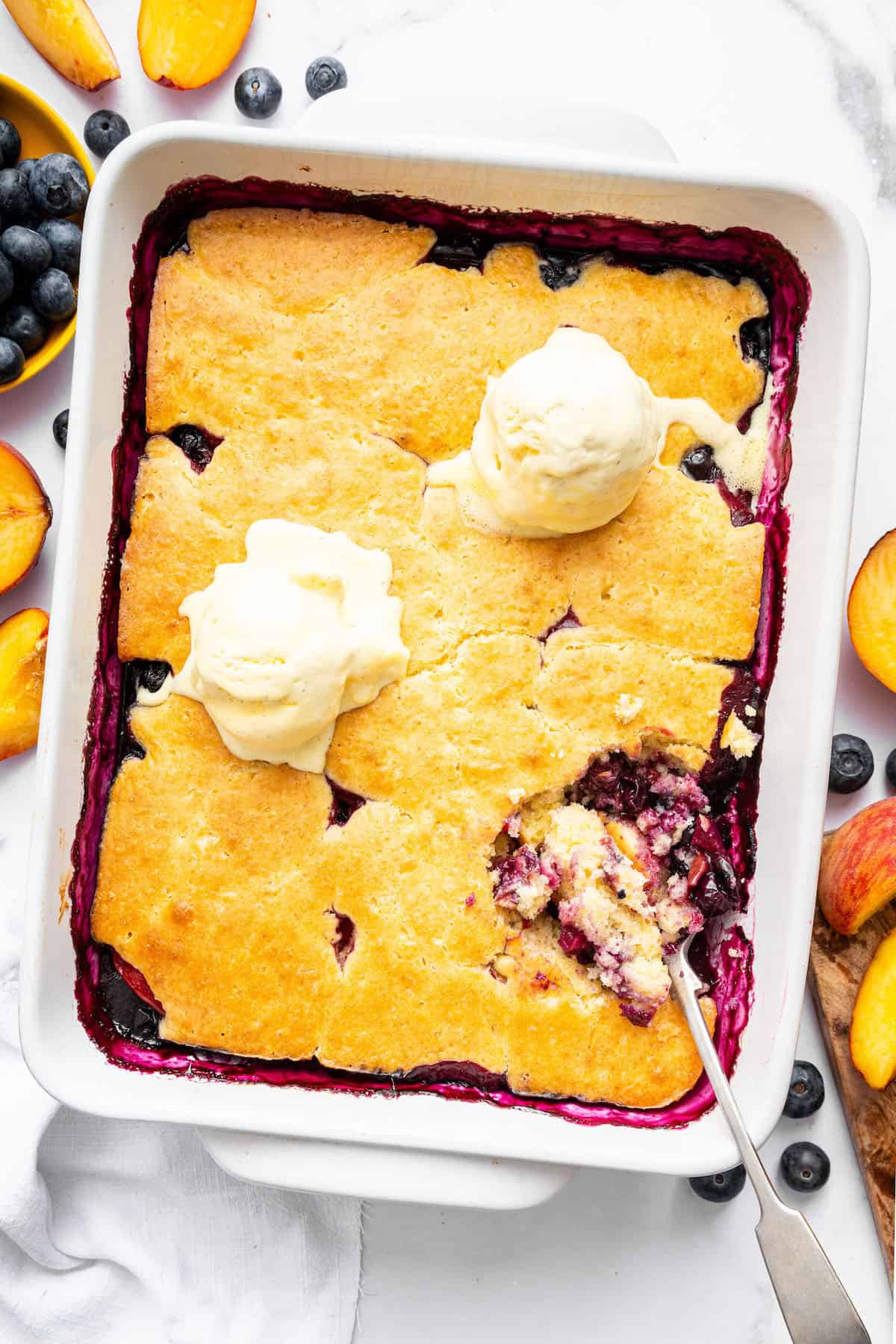 This blueberry peach cobbler recipe pairs two summer favorites in a jammy filling that’s baked with a buttery, flaky topping. So easy! Overhead view of blueberry peach cobbler in baking dish with scoops of vanilla ice cream and spoon