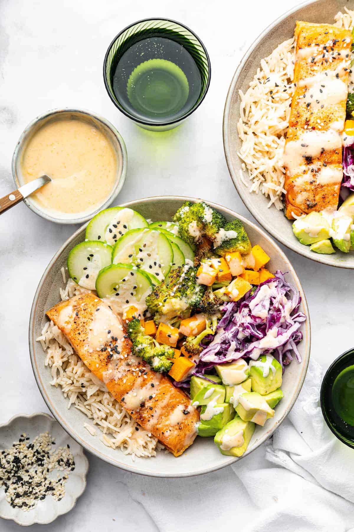 With flaky salmon, grains and veggies, this Salmon Power Bowl recipe is an easy meal prep option that will fuel you through your day! Overhead view of two salmon power bowls with small bowl of sauce, plate of sesame seeds, and water glasses