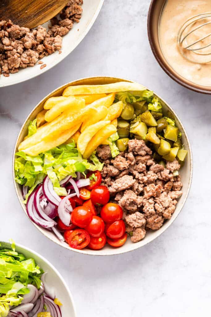 This Burger Bowl recipe gives you your burger fix, without the bun! It’s a fun protein-packed meal with crispy fries and your fave toppings. Burger bowl with ground beef, pickles, fries, lettuce, onion, and tomatoes.