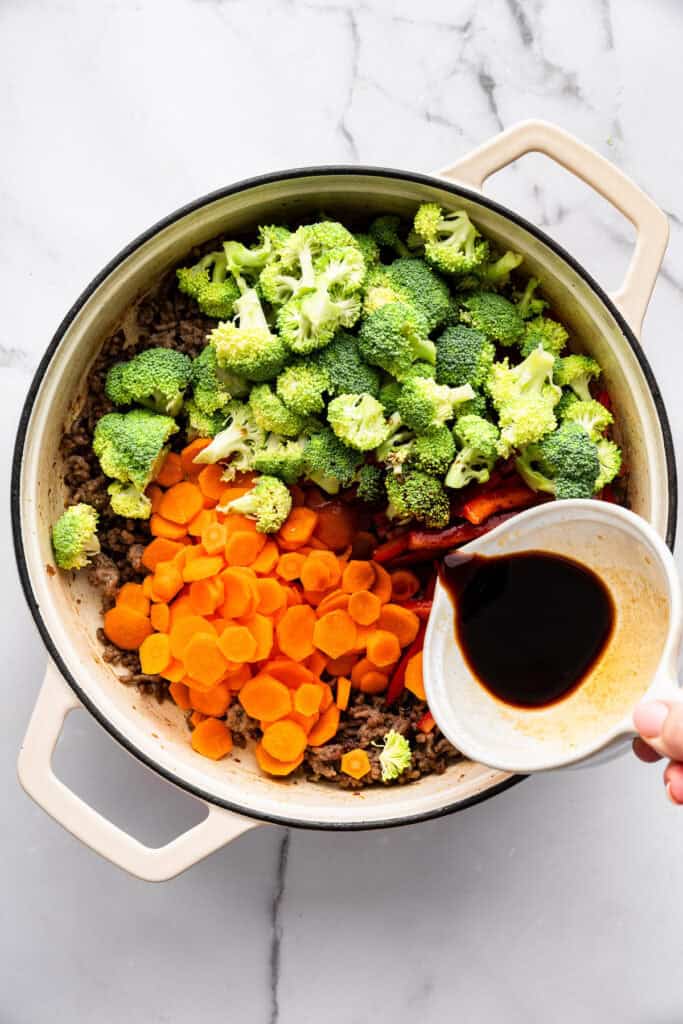 Springy ramen noodles are combined with ground beef and veggies to make this super easy one pan Beef Ramen recipe. Done in 25 minutes! Pouring sauce into pan with sliced carrots and broccoli.