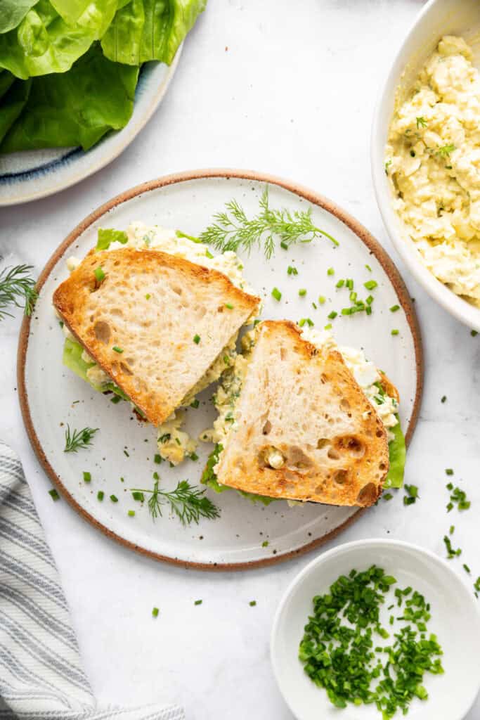 This classic egg salad sandwich on toasty sourdough is tasty and satisfying. It's perfect for an easy lunch that's ready in just 5 minutes! Egg salad sandwich cut in half on plate with fresh herbs.