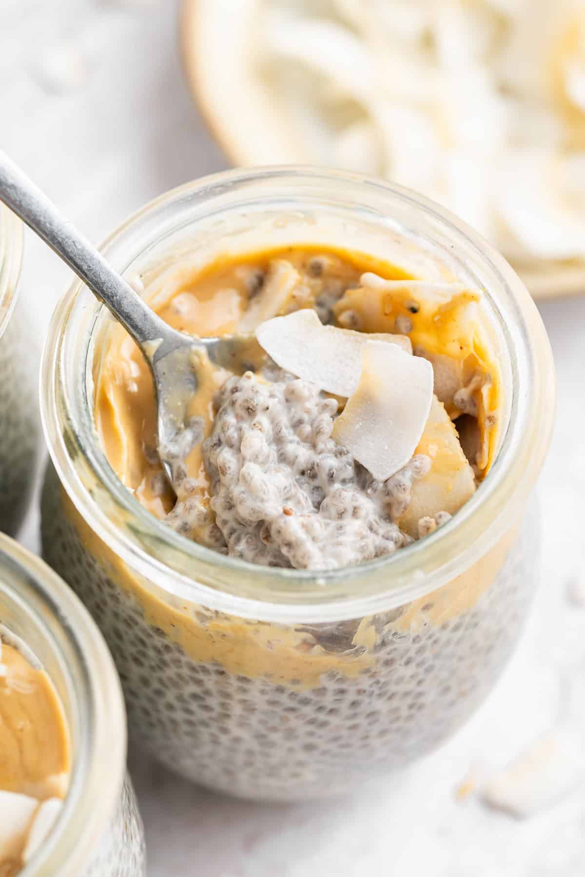 A spoon diving into a jar of coconut chia pudding topped with cashew butter and coconut flakes.