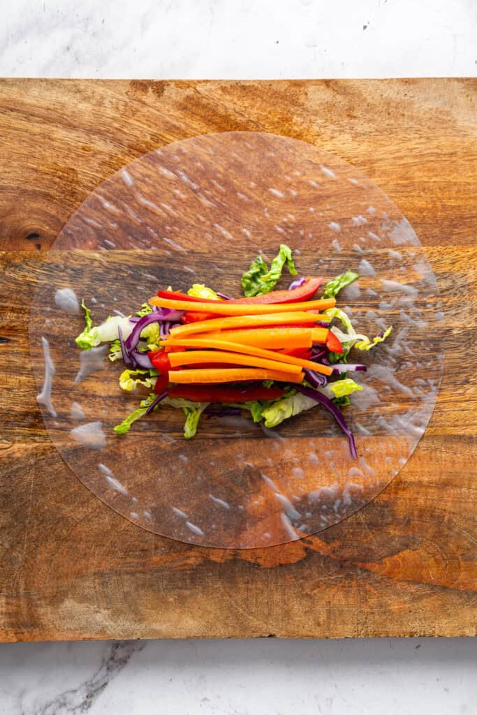 On warm days, this summer rolls recipe is a must. Crisp veggies and avocado are wrapped in rice paper for a refreshing, no-cook meal. A hydrated rice paper wrapper with shredded romaine, red cabbage, bell pepper slices, and carrot slices on top.