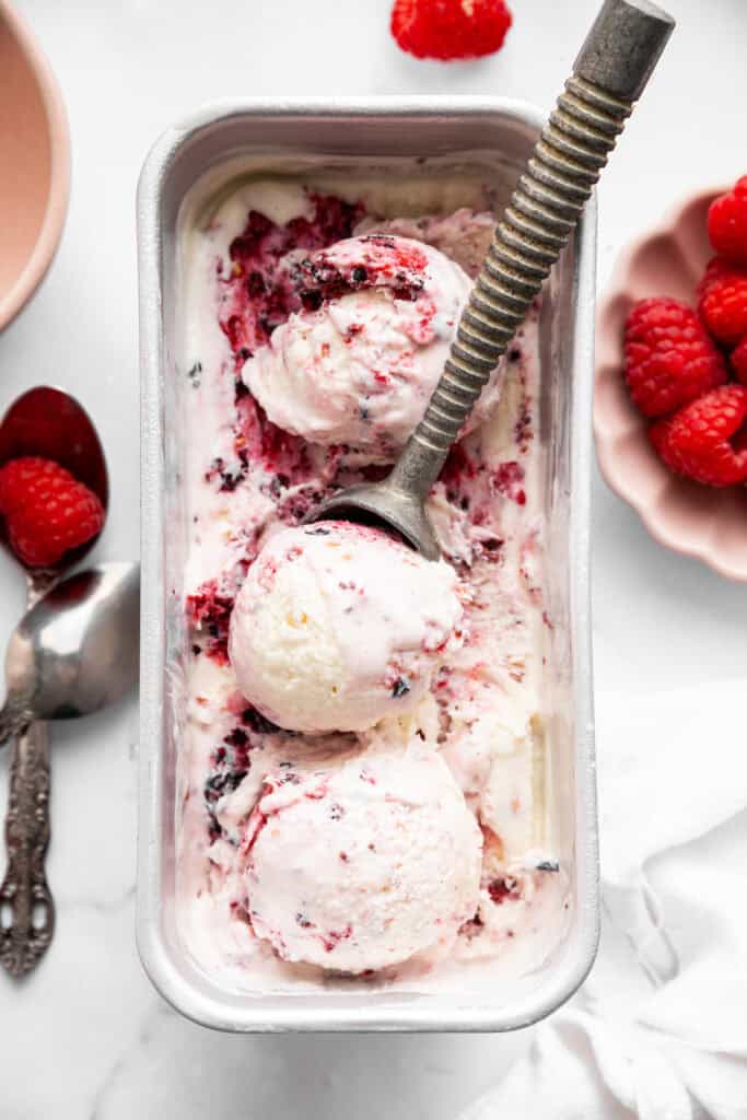 This Greek yogurt ice cream is smooth, creamy, packed with protein, and lightly sweetened with honey and a colorful swirl of fresh berries. An ice cream scooper diving into a tub of Greek yogurt ice cream with a berry swirl.