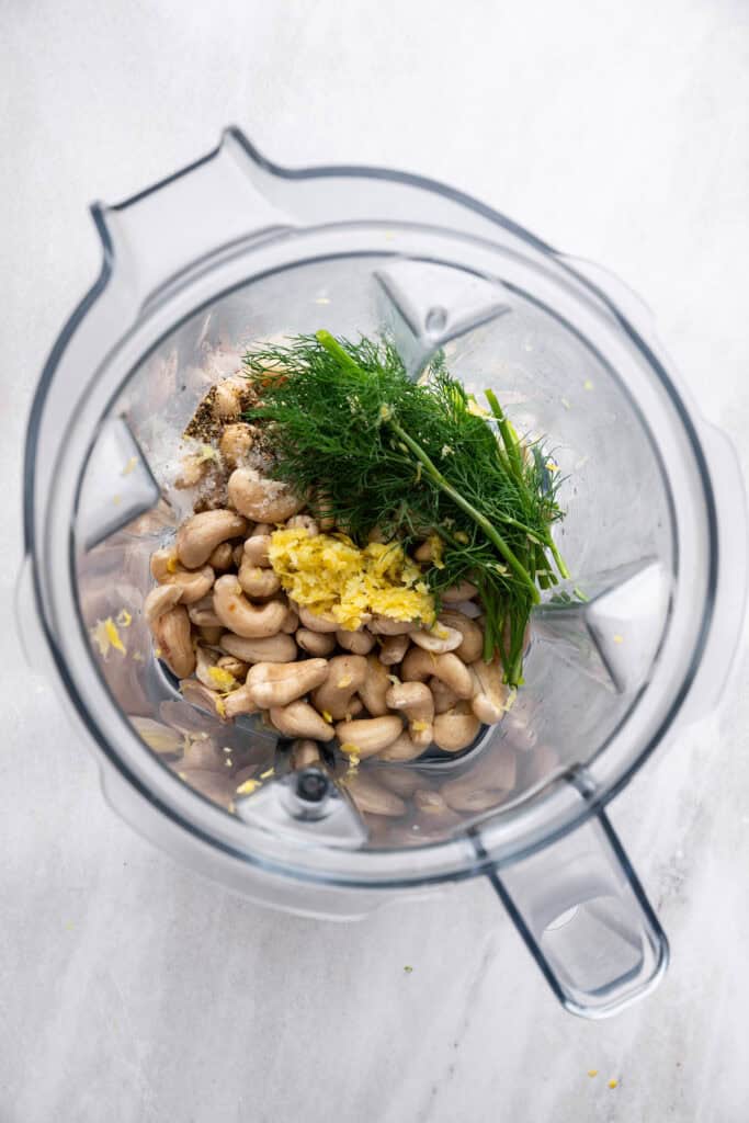 Adding soaked cashews, lemon juice, lemon zest, fresh dill, water, salt, and pepper to a blender.